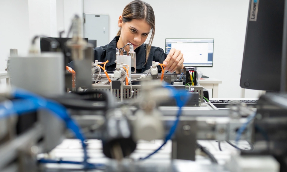 Engineer working on automated electronic testing equipment, adjusting components and connections while monitoring data on a computer in a laboratory environment.