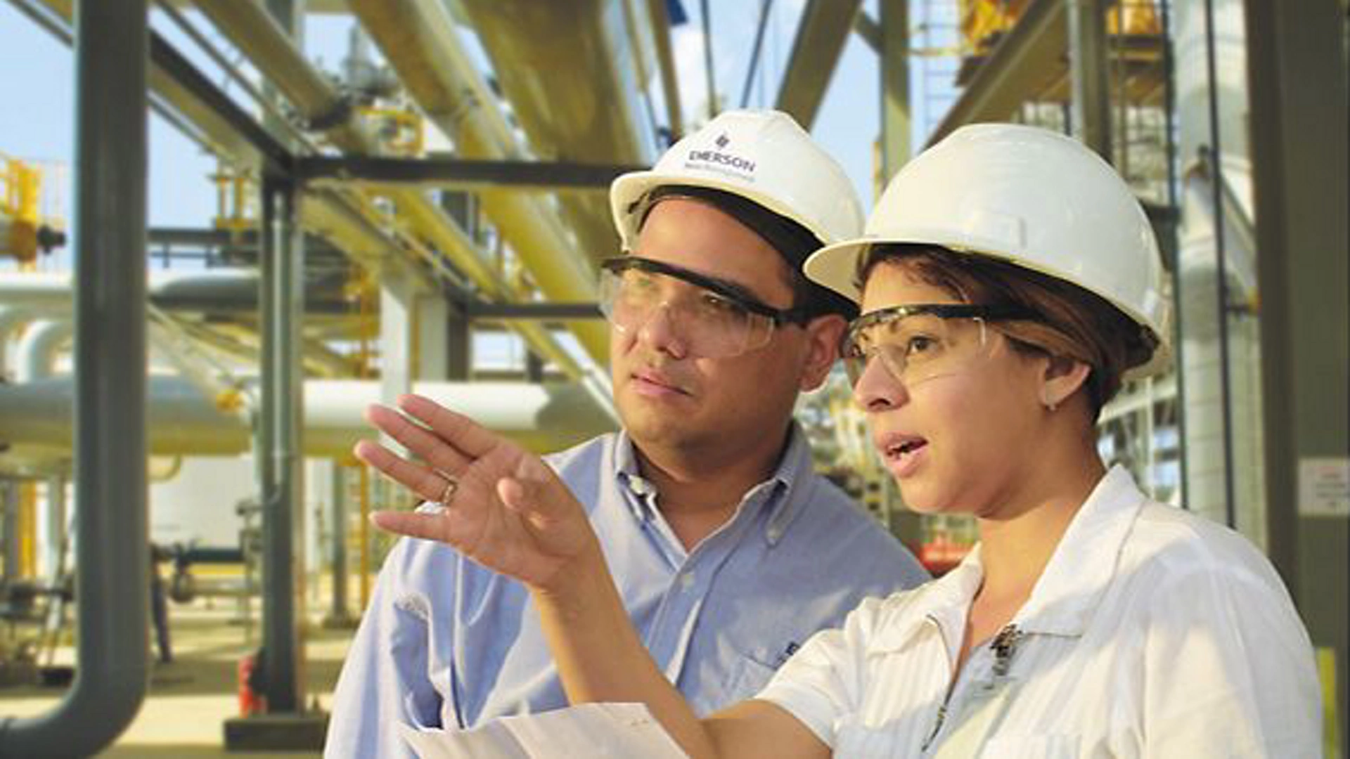 Two workers wearing hard hats are engaged in a discussion at an industrial facility with visible pipelines and structural elements in the background. One worker is gesturing while holding a document, indicating a collaborative or instructional moment. The setting suggests a focus on engineering, construction, or industrial operations.
