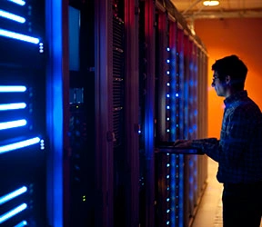 The interior of a modern server room in a data center.  An IT engineer is busy configuring the servers. The room is dark, but the servers themselves are lit.  The servers at the left side are lit in blue, while the ones at the other end are lit by an orange light source coming from the right side.