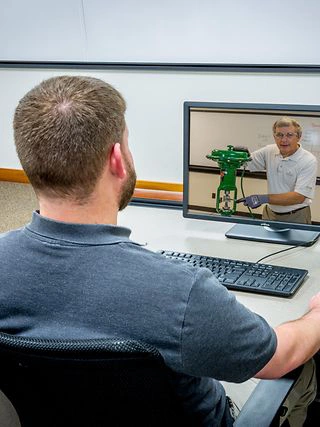 A man sits at a desk in an office setting, viewing an instructional video on a computer screen. The video features a person demonstrating a green mechanical device. The environment includes a whiteboard in the background and standard office furniture. The focus is on learning and technical demonstration.