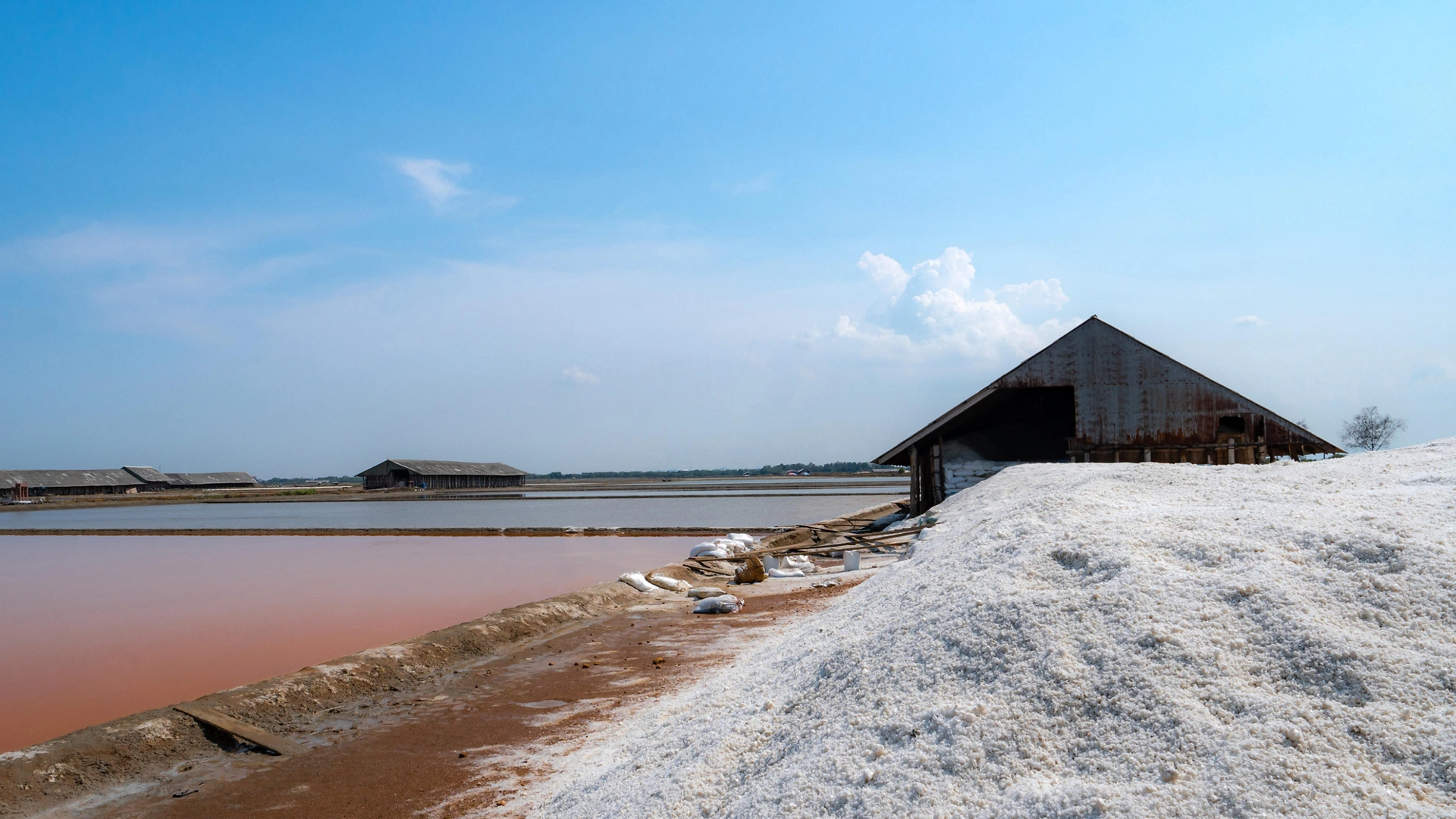 Landscape of brine salt farm with blue sky on sunny day. Salt warehouse. Pile of organic sea salt. Raw material of salt industrial. Sodium chloride mineral. Evaporation of sea water. Iodine.